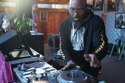 DJ Cee White commands the turntables during the Your Sunday Best season opener at White Owl Social Club in Portland's vibrant music scene. Warm afternoon light filters through the intimate venue as the bearded performer, wearing a black hoodie with yellow checkerboard details, manipulates vinyl records with practiced precision. The cozy club atmosphere, complete with potted plants and framed artwork on wood-paneled walls, creates the perfect backdrop for this daytime celebration of hip-hop culture.