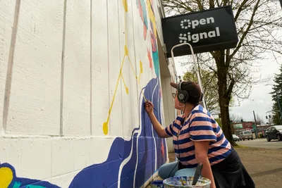 Maria Rodriguez, known as Sparkykneecap, meticulously applies blue paint to a vibrant mural on the white corrugated exterior of Open Signal in Portland's Eliot neighborhood. The Mexican-American artist, wearing headphones and a striped shirt, works from a ladder while creating the commissioned piece titled "Let's Talk" for the Regional Arts & Culture Council. Soft spring light filters through bare tree branches, illuminating the evolving artwork that explores themes of identity and cultural connection through bold geometric shapes and vivid colors.