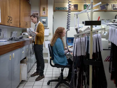 Two film lab technicians work in the intimate, equipment-filled processing room at Blue Moon Camera & Machine in Portland's Saint Johns neighborhood. A woman with curly red hair sits at a professional film processing station while her colleague in a mustard yellow cardigan examines negatives by the counter. The warm fluorescent lighting illuminates vintage processing equipment, coiled cords, and the organized chaos of a working analog photography lab.