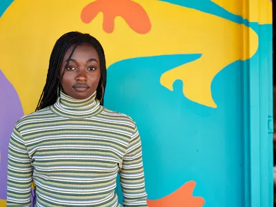 Naomi Likayi stands confidently before her completed mural in Portland's Alberta Arts District, her long braids framing her face as she poses in a striped turtleneck. The explosive backdrop features sweeping organic forms in brilliant turquoise, sunshine yellow, and coral orange that dance across the wall behind her. Natural light illuminates both artist and artwork, creating a harmonious dialogue between creator and creation in this intimate moment of artistic completion.