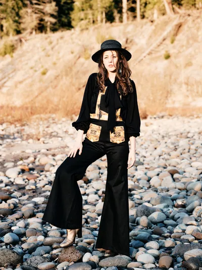 A young woman stands contemplatively on a riverbed of weathered stones along the Floodplain Trail in Multnomah County, Oregon. She wears a striking black ensemble with metallic embellishments and a wide-brimmed hat, her flowing hair catching the golden hour light. The composition captures her against the textured landscape of river rocks and distant hillsides, creating an atmospheric portrait that blends fashion with the raw beauty of the Pacific Northwest wilderness.