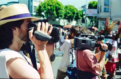 A bearded man in a woven straw hat with purple band operates a handheld Sony video camera in the foreground, while another person with blonde hair films behind him during a community demonstration. The scene unfolds on a sun-drenched Mission District street, with leafy green trees and urban buildings creating a backdrop of neighborhood activism. The warm afternoon light captures the grassroots nature of residents documenting their own struggle against gentrification.