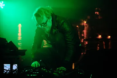 Electronic musician Eli Savage, performing as ellipsism, hunches intensely over his synthesizer setup in the emerald glow of stage lighting at Dante's in Portland. The atmospheric green wash transforms the intimate venue into an otherworldly sonic laboratory, while warm amber lights punctuate the darkness behind him. His focused posture and tousled hair capture the raw concentration of live electronic performance, as cables and equipment sprawl across the stage like digital arteries feeding his sonic creation.