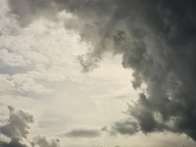 Brooding cumulonimbus clouds mass in pewter and charcoal tones above Portland's Central Eastside district, their heavy forms caught in dramatic chiaroscuro against patches of luminous sky. The monochromatic palette emphasizes the raw power of the approaching weather system, with billowing cloud formations creating a sense of impending atmospheric drama over the city's industrial corridor. Light breaks through in ethereal beams, casting the entire skyscape in theatrical contrast between shadow and illumination.