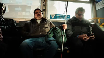 Golden morning light streams through the windows of a TriMet bus as it carries commuters through Northeast Portland's historic Eliot district. A passenger with a white cane sits peacefully in the priority seating area, while another commuter checks his phone, both bundled against the Pacific Northwest chill in dark winter jackets. The warm interior glow contrasts with the pale overcast sky visible through rain-spotted windows, capturing the quiet rhythm of urban transit accessibility.