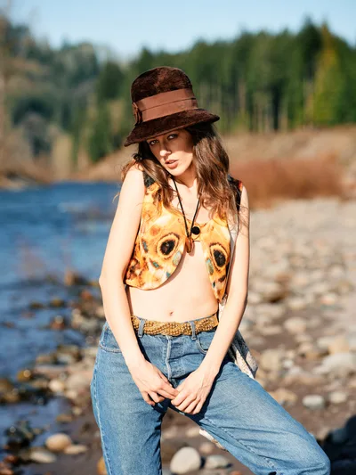 A young woman in bohemian attire poses confidently along the rocky riverbed of the Columbia River in Oregon's Floodplain Trail. She wears a distinctive owl-printed crop top, vintage denim, and a brown felt hat with leather band, creating a free-spirited aesthetic. Golden hour light illuminates her face while the river's blue waters and forested hills of Multnomah County create a dreamy backdrop. The composition captures the essence of Pacific Northwest outdoor lifestyle and artistic self-expression.