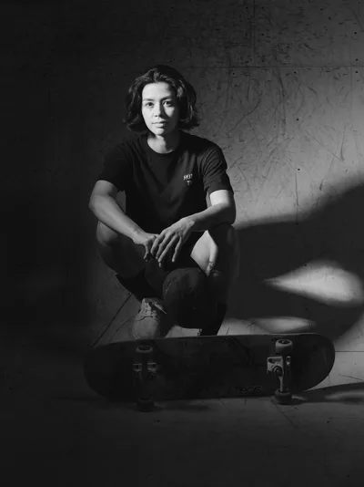 Pro skateboarder Christiana Means sits in contemplative repose against weathered concrete walls at Stronger Skatepark in Milwaukie, Oregon. The monochromatic portrait captures her relaxed posture with arms draped over bent knees, her skateboard positioned prominently in the foreground. Dramatic lighting carves shadows across the textured surfaces, creating an intimate study of athletic grace in an urban sanctuary.