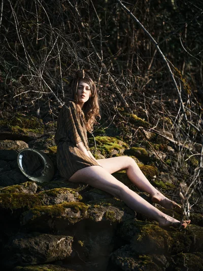 A young woman with long flowing hair sits barefoot on moss-covered rocks along Oregon's Floodplain Trail, bathed in golden hour light that filters through the dense woodland behind her. She wears an earth-toned jacket and holds what appears to be a hat, gazing contemplatively into the distance. The composition captures the intimate relationship between human presence and the ancient, moss-draped stone formations characteristic of the Pacific Northwest's riverbed environments.