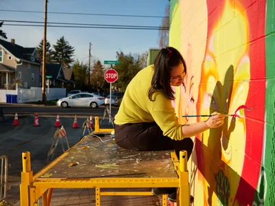 Multidisciplinary artist Villatose meticulously applies brushstrokes to her mural 'Jardín Cubana,' depicting three female figures in vibrant colors on Northeast Alberta Street in Portland's celebrated arts district. Golden hour light bathes the scene as she works from a yellow scaffolding platform, her concentrated expression captured against the vivid spectrum of colors cascading across the wall. The residential neighborhood backdrop, complete with power lines and a stop sign, frames this moment of creative transformation in one of Portland's most dynamic cultural corridors.
