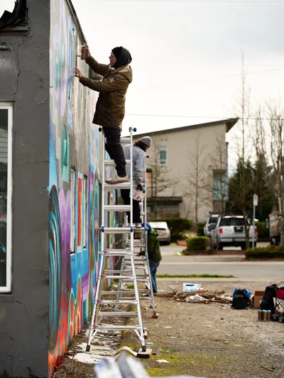 A dedicated muralist in a brown winter jacket stretches across a ladder to add vibrant brushstrokes to an evolving street art piece on a gray building wall. The scene captures the intimate process of community art creation on an overcast Northeast Portland afternoon, with residential buildings and bare winter trees providing a quiet suburban backdrop. Paint supplies and materials scatter across the ground below, while a second artist works nearby, testament to the collaborative spirit of neighborhood beautification projects.