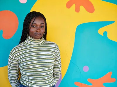 Naomi Likayi stands confidently before her completed abstract mural in Portland's Alberta Arts District, her dark braids framing her face as she wears a striped turtleneck. The dynamic backdrop features flowing organic shapes in brilliant turquoise, golden yellow, coral orange, and soft pink that seem to dance around her figure. The composition captures both the artist's quiet pride and the exuberant energy of her colorful creation, embodying the creative spirit of Northeast Portland's vibrant arts community.