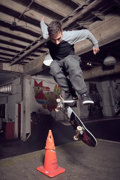 A young skateboarder launches into a gravity-defying ollie over an orange traffic cone in the raw, industrial interior of Commonwealth Skateboarding in Portland's Buckman neighborhood. The weathered wooden ceiling beams and graffiti-tagged concrete walls create a gritty urban canvas, while dramatic lighting captures the precise moment of aerial suspension. His focused expression and athletic form demonstrate the technical mastery required for this fundamental skateboarding trick.
