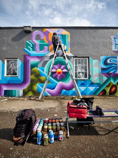 A bundled figure in dark clothing balances on an aluminum ladder, spray painting a dynamic geometric mural across the weathered gray facade of a Northeast Portland building. The kaleidoscopic artwork explodes with interlocking shapes in purple, teal, orange, and electric blue, featuring dimensional letters and a radiating mandala-like centerpiece. At ground level, an arsenal of spray paint cans stands organized in neat rows alongside the artist's equipment, creating a tableau of urban creativity beneath Oregon's characteristic overcast sky.