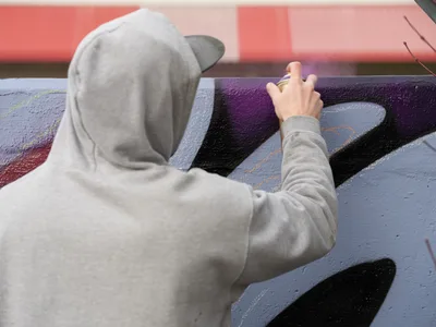 Travis Fields, known as Campographic, works intently on a vibrant mural at the Clay Creative Building in southeast Portland's Hosford-Abernethy neighborhood. Wearing a gray hooded sweatshirt, the artist reaches toward bold purple and black geometric forms painted on the concrete wall. The composition captures the focused concentration of street art creation, with warm coral-pink tones from adjacent surfaces creating atmospheric depth against the cool wall colors.