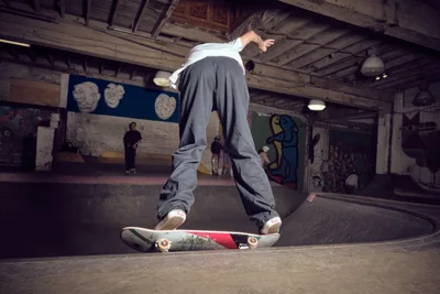 A skateboarder navigates the curved concrete bowl at Commonwealth Skateboarding in Portland's Buckman neighborhood, his baggy grey pants and white shirt creating dynamic lines against the weathered walls. The industrial space hums with underground energy, its exposed wooden beams and graffiti-adorned walls bearing witness to countless sessions. Onlookers gather in the shadows while pendant lights cast a warm glow over this temple of skateboard culture.