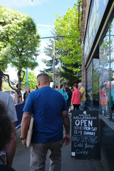 A diverse crowd of music enthusiasts gathers on the tree-lined sidewalk outside Music Millennium on East Burnside Street in Portland's Kerns neighborhood. The iconic independent record store's storefront displays vibrant album artwork in its windows, while a prominent chalkboard sign announces operating hours and cold beverages available. Dappled afternoon sunlight filters through the urban canopy, casting a warm glow over the patient queue of customers waiting to enter this beloved musical institution.