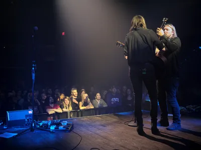 Emma Richardson and Russell Marsden of Band of Skulls perform during their 10 Year Anniversary Tour at Webster Hall in Manhattan's East Village. The duo stands center stage bathed in dramatic stage lighting, with Richardson holding her guitar while Marsden faces away from the camera. The intimate venue setting captures the engaged audience seated at tables in the foreground, creating a sense of connection between performers and fans in this historic New York music venue.