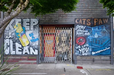 Weathered wooden storefronts along Southeast Division Street display contrasting pandemic-era murals beneath overhanging maple branches. The left mural promotes hygiene with "KEEP it CLEAN" messaging and sanitizer imagery, while the right celebrates local resilience with "CAT'S PAW Stay Strong Portland We Love You" in bold lettering. A security grate protects the central entrance at address 3565, creating an urban tableau of community solidarity and public health awareness.