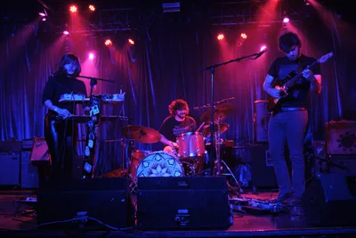 Experimental band Sun Blood Stories commands the intimate stage at Dante's in downtown Portland, bathed in crimson and violet stage lights that pierce through the smoky atmosphere. The three-piece band from Boise, Idaho—featuring a keyboardist, drummer, and guitarist—creates their mesmerizing modern-day opera sound amid the venue's signature industrial backdrop of exposed metal rigging and curtained walls. The moody lighting casts dramatic shadows while highlighting the concentrated energy of the musicians mid-performance.