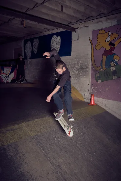 A skateboarder executes a trick in the dimly lit basement of Commonwealth Skateboarding in Portland's Buckman neighborhood. The concrete space is adorned with colorful cartoon murals, including character faces on blue backgrounds and a Simpsons-style figure. Flash photography captures the raw energy of the underground skate scene, with another figure visible in the shadowy background as wooden ceiling beams frame the gritty urban sanctuary.