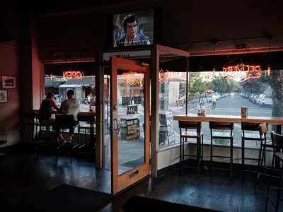 The warm amber glow of neon signs illuminates the interior of Straight From New York Pizza on Portland's Hawthorne Boulevard, where two patrons sit at a dark wooden table in the foreground. Through expansive glass windows, the tree-lined street comes alive with evening pedestrians and cyclists, while a vintage television screen casts additional light from above. The scene captures the quintessential neighborhood pizzeria atmosphere where indoor comfort meets the vibrant pulse of Southeast Portland's cultural corridor.