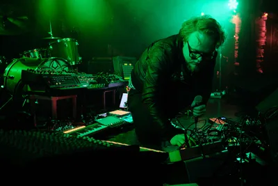 Electronic musician Eli Savage, performing as Ellipsism, leans intently over his synthesizer setup during a live performance at RXFest 2019. Bathed in vivid emerald stage lighting that transforms the intimate venue into an otherworldly sonic laboratory, the bearded artist manipulates a dense array of electronic equipment while gripping a microphone. The atmospheric lighting casts deep shadows across Dante's club in Portland's downtown district, creating a moody tableau where technology and artistry converge in the underground music scene.