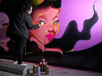Multidisciplinary artist Vilatose meticulously applies paint to a striking feminine portrait rendered in electric pinks and purples against the concrete wall of Clay Creative's parking garage in Portland's Hosford-Abernethy neighborhood. The masked artist, clad in all black, works with focused precision as an arsenal of spray paint cans sits ready on the pavement below. The mural depicts a sultry female figure with dramatic eyeliner and flowing dark hair, her image emerging from shadows into brilliant magenta light.
