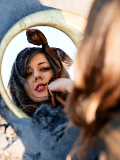 A contemplative moment captured through a vintage hand mirror held along the Floodplain Trail in Multnomah County, Oregon. The reflection reveals a young woman with dark hair adorned with a tortoiseshell headband, her red lips and gentle expression framed by the mirror's weathered brass edge. Warm golden sunlight filters through the scene, creating an intimate interplay between the reflected subject and the blurred riverbed environment surrounding the trail.