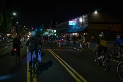 Dozens of cyclists converge on Southeast Division Street during Portland's Drop Out Prom Ride, part of the city's annual Pedalpalooza celebration. The nocturnal scene pulses with red bicycle lights scattered across the asphalt like urban fireflies, while Tom's Bar's neon sign casts an electric blue glow over the assembled riders. Helmeted participants in casual attire cluster in the street and on sidewalks, their bicycles creating an impromptu street festival atmosphere beneath the warm amber streetlights of this Richmond neighborhood intersection.