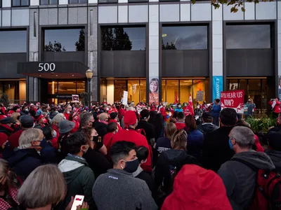 A dense crowd of healthcare workers, predominantly wearing red jackets and face masks, congregates outside the Kaiser Permanente Tower at 500 Northeast Multnomah Street in Portland's Lloyd District. The sea of protesters fills the frame from foreground to the building's glass facade, creating a powerful visual of collective action. Protest signs punctuate the crowd, including one reading 'CAN YOU HEAR US NOW?' while the building's modernist architecture looms behind them, its illuminated interior contrasting with the gathering dusk outside.