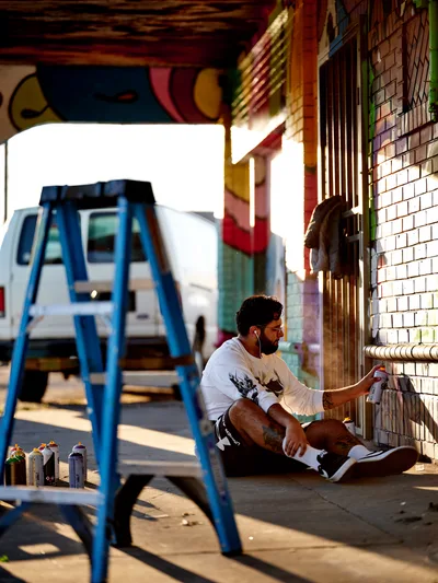 Muralist Heysus sits cross-legged on a sidewalk in Portland's Alberta district, spray painting intricate details on a colorful brick wall mural. Golden hour light streams through the urban scene, casting warm shadows from a blue metal stepladder in the foreground while a white RV sits parked in the soft-focus background. The artist works with focused concentration, earbuds in, surrounded by scattered spray paint cans on the concrete.