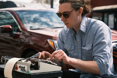 Till Gwinn, a local street poet, concentrates intently as he types on a vintage white typewriter at his outdoor workspace on Northeast Alberta Street in Portland. Wearing a pale blue button-down shirt and tortoiseshell sunglasses, he sits with legs spread apart while putting finishing touches on a custom poem for a waiting customer. The warm afternoon light illuminates his focused expression while a burgundy pickup truck provides an atmospheric backdrop to this quintessential Portland street scene.