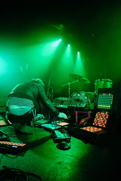 Electronic musician Eli Savage performs as Ellipsism at RX Fest 2019, hunched over an intricate array of synthesizers and drum machines at Portland's iconic Dante's venue. Piercing emerald spotlights cut through the smoke-filled darkness, casting an otherworldly glow over the densely packed performance setup. The artist's silhouette disappears into the maze of glowing control panels and grid controllers that sprawl across the stage floor, creating an immersive sonic laboratory bathed in electric green luminescence.