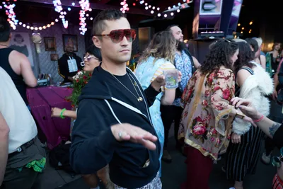 A man in tortoiseshell aviators and a black tracksuit gestures mid-conversation while holding a drink at Your Sunday Best, a vibrant summer day party at Portland's White Owl Social Club. The intimate venue pulses with string lights and eclectic wall art as diverse partygoers in floral prints and casual attire mingle in the warm, bokeh-lit atmosphere. The scene captures the relaxed yet energetic spirit of Portland's underground music scene, where afternoon revelry unfolds beneath cascading fairy lights.