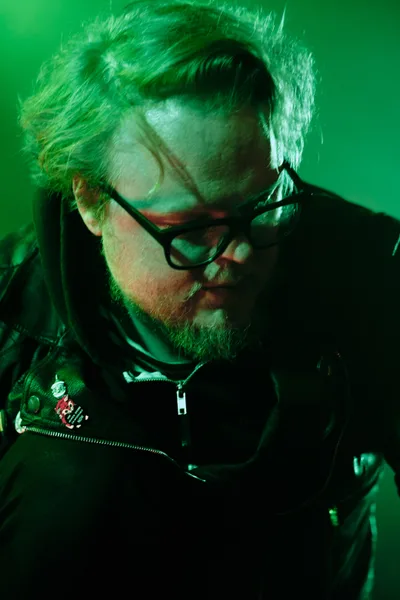 Electronic musician Eli Savage, performing as Ellipsism, becomes lost in concentration during his live set at RXFest 2019. Bathed in atmospheric green lighting at Dante's venue in Portland, Oregon, his silver hair catches the theatrical glow as he manipulates his equipment. The intimate portrait captures the artist's complete immersion in his craft, black-rimmed glasses reflecting the stage lights while patches on his dark jacket hint at his underground aesthetic.