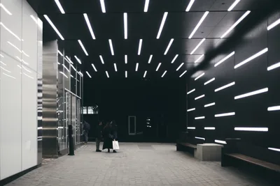 Dramatic linear LED lights pierce through the darkness of a modern transit station, creating a mesmerizing starburst pattern that radiates from a central vanishing point. Commuters move through this stark architectural space where gleaming steel elevator banks contrast against the deep shadows, their silhouettes dwarfed by the monumental scale of the underground concourse. The geometric precision of the lighting installation transforms utilitarian infrastructure into a cathedral of contemporary urban design, casting sharp white beams that slice through the cavernous darkness.