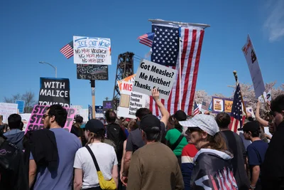 Demonstrators gather beneath the clear blue Oregon sky on Northwest Naito Parkway in Portland's historic Old Town district, their voices rising in unified dissent. American flags flutter alongside handmade signs declaring messages against government overreach, including prominent placards reading "ICE OUT NOW!" and "Got Measles? Me Neither!" The crowd stretches into the distance, a sea of diverse faces and colorful protest signs framed by the industrial architecture of Portland's waterfront. Spring light illuminates the passionate assembly as citizens exercise their constitutional right to peaceful protest.