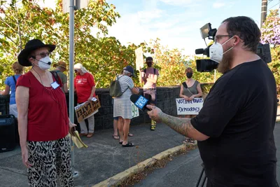 A tattooed television cameraman from KOIN Channel 6 interviews a climate activist wearing a red top and black hat outside Representative Kurt Schrader's Oregon City office. The woman stands confidently before the camera while fellow demonstrators gather behind her, their handmade signs advocating for voting rights and democracy visible in the dappled afternoon light. The scene unfolds on McLoughlin Promenade, where autumn foliage frames this grassroots moment of civic engagement.