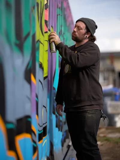 A bearded artist in a charcoal beanie and dark hoodie applies spray paint to a vivid mural featuring electric greens, purples, and blues on a wall in Portland's Alberta district. His concentrated expression and deliberate movements capture the intimate process of urban art creation. The blurred cityscape behind him emphasizes his focused connection with the colorful canvas before him, while natural daylight illuminates both artist and artwork in this candid moment of creative expression.