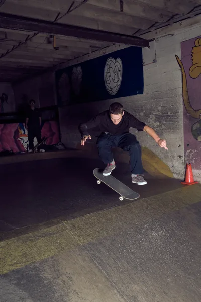 A skateboarder navigates the dimly lit basement of Commonwealth Skateboarding in Portland's Buckman neighborhood, his form silhouetted against vibrant graffiti murals. The industrial concrete space, marked by exposed ceiling beams and painted brick walls, creates an intimate underground atmosphere where street art and skateboarding culture converge. Harsh artificial lighting casts dramatic shadows as the athlete maintains balance mid-trick, while a solitary traffic cone punctuates the raw urban setting.