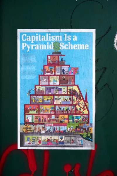 A weathered poster declaring 'Capitalism Is a Pyramid Scheme' adheres to a dark green wall in Portland's Concordia neighborhood, its edges worn from exposure. The detailed illustration depicts a multi-tiered pyramid structure with colorful cross-sections showing different socioeconomic levels, from luxury penthouses crowned with dollar signs at the top to cramped basement conditions at the bottom. Red graffiti marks streak beneath the poster, creating a stark contrast against the emerald backdrop while emphasizing the raw, urgent nature of this pandemic-era street commentary.
