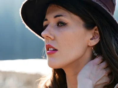 A young woman gazes contemplatively into the distance, her profile illuminated by warm golden light filtering through her wide-brimmed hat. The intimate portrait captures her wearing delicate chain earrings and a soft pink garment, with the dreamy bokeh of Oregon's riverbed trail creating an ethereal backdrop. The gentle lighting emphasizes the natural texture of her skin and the subtle gradation from shadow to light across her features.