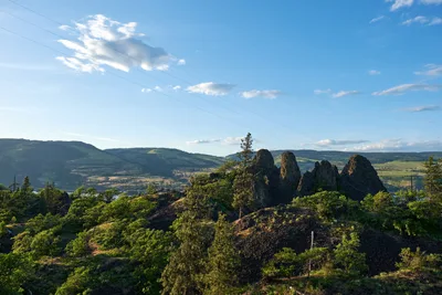 Jagged basalt rock formations pierce through verdant vegetation on Oregon's Memaloose Plateau, creating a dramatic foreground against the sweeping Columbia River Gorge valley. Power lines traverse the azure sky dotted with white clouds, while rolling hills extend toward the horizon in layers of green and gold. The afternoon light illuminates the volcanic remnants and lush Pacific Northwest landscape, capturing the raw geological beauty where ancient lava flows carved this iconic terrain.